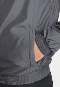 Gray windbreaker jacket with elastic cuffs and waistband. Features side pockets and a smooth, water-resistant texture.