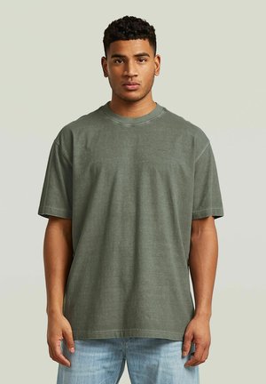 Jeune homme aux cheveux courts et bouclés portant un t-shirt vert uni oversize et un jean bleu clair, debout devant un fond neutre.
