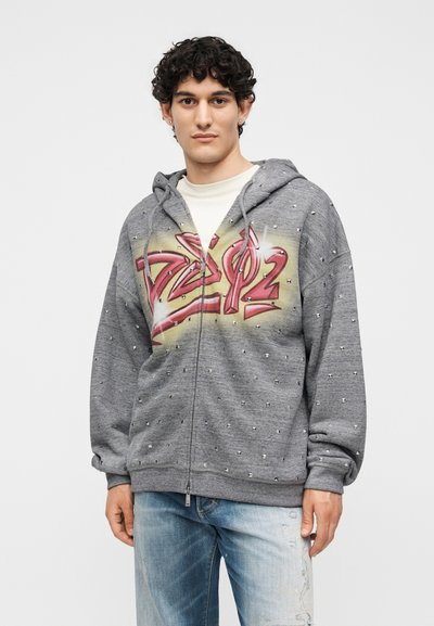 Νέος άνδρας με γκρι φούτερ με φερμουάρ και διακοσμητικές τρουκς, με κόκκινο κείμενο σε στυλ γκράφιτι, συνδυασμένο με ανοιχτά μπλε τζιν με φθορές.