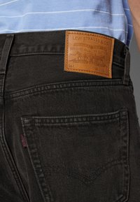 Gros plan sur la poche arrière noire d'un jean Levi's et l'étiquette de marque en cuir marron à la taille, porté avec une chemise rayée bleu clair.