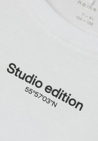 Hvit t-skjorte med rund hals; har svart tekst "Studio edition 55°57'03'N" på forsiden. Mykt bomullsmateriale med en glatt tekstur.