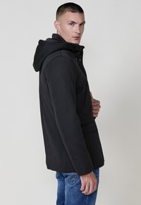 Giacca nera con cappuccio realizzata in tessuto testurizzato; presenta una tasca con zip sul petto e tasche laterali; indossata sopra jeans blu.