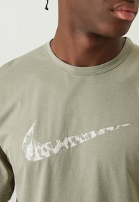 Oliwkowa bawełniana koszulka z białym logo Nike w kształcie łuku z delikatnym efektem teksturowania. Klasyczny krój z okrągłym dekoltem. Krótkie rękawy.