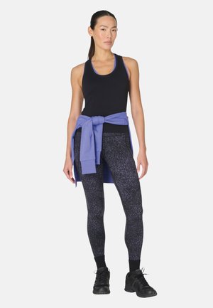 Frau steht, trägt ein schwarzes Tanktop, schwarz-weiß gesprenkelte Leggings, schwarze Schuhe und einen blauen Pullover, der um die Taille gebunden ist.