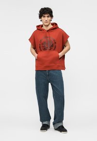 Jonge man staand met handen in de zakken van zijn hoodie, draagend een rood mouwloos hoodieshirt met grafische tekst, losse blauwe spijkerbroek en zwarte sneakers.