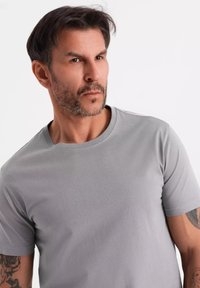 T-shirt in cotone grigio con colletto a girocollo e maniche corte, caratterizzato da una texture liscia e una vestibilità comoda. Tatuaggi visibili sul braccio.
