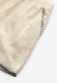Pantaloni corti crema con un motivo testurizzato di foglie di palma. Vita elasticizzata e cuciture nere a contrasto lungo la cucitura. Materiale leggero.