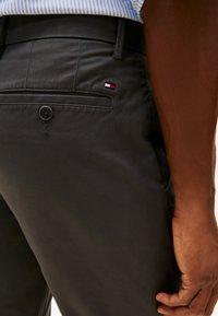 Calças cinzentas escuras com uma textura suave, apresentando um bolso traseiro e um pequeno emblema com as cores vermelho, branco e azul. Detalhe de fecho com botão visível.