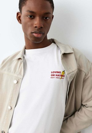 Jeune homme portant une veste beige sur une chemise blanche avec un texte rouge « AIMER OU DÉTESTER SALÉ • FRAIS • ÉPICÉ » et un petit graphique de piment.