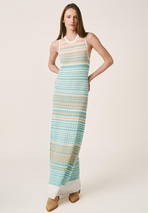 Mujer de pie con las manos detrás de la espalda, vistiendo un vestido de punto sin mangas y largo con rayas onduladas horizontales en tonos teal, beige y blanco.