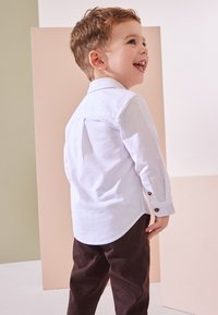 Camicia bianca a maniche lunghe con dettaglio a bottoni, abbinata a pantaloni marrone scuro, indossata da un bambino visto di spalle su uno sfondo neutro.