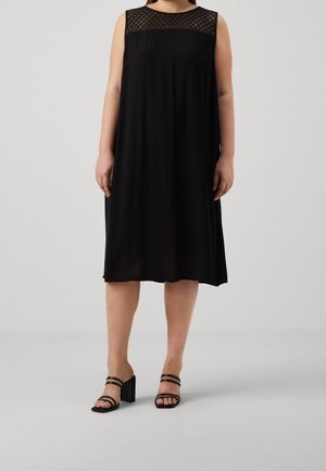 Abito nero senza maniche realizzato in tessuto fluido, con un pannello in pizzo testurizzato sullo scollo. La modella indossa sandali neri con tacco.
