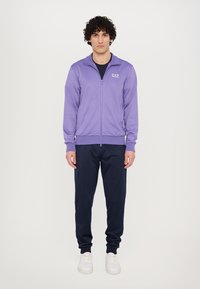 Violets krāsas zip-up sporta jaka ar stāvo apkakli, logo uz krūts, apvienota ar tumši ziliem sporta biksēm un baltām kedām. Gluda, viegla auduma.