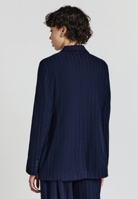 Blazer a righe sottili blu con vestibilità su misura, lapel a punta e dettaglio con tre bottoni ai polsini. Texture liscia e silhouette strutturata.