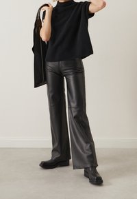 Top de punto negro con cuello alto, combinado con pantalones de cuero negros acampanados. La modelo sostiene un gran bolso negro y lleva botas gruesas negras.