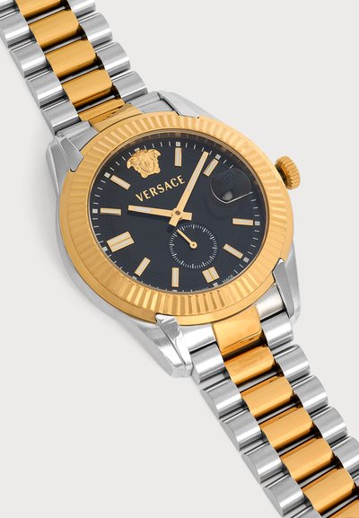 Montre de luxe avec cadran noir, lunette dorée et bracelet métallique bicolore argent et or, avec logo Versace et loupe pour la date.