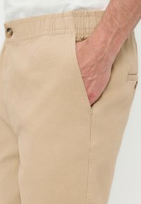 Χέρι στην τσέπη παντελονιού με μπεζ ελαστική μέση και λεπτομέρεια κουμπιού στην πλευρική τσέπη, σε ουδέτερο φόντο.