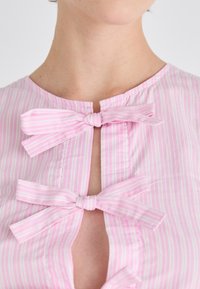 Camicetta rosa chiaro con strisce bianche verticali, caratterizzata da una chiusura a collo a chiave fissata da tre fiocchi legati con tessuto coordinato.