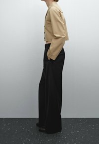 Person standing in beige cropped jacket and loose black pants with hands in pockets against plain wall.