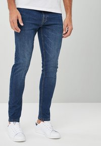 Jeans de denim ajustados de color azul oscuro, con un diseño clásico de cinco bolsillos y detalles de costura sutiles, combinados con zapatillas blancas.