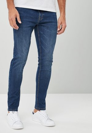 Jeans de denim ajustados de color azul oscuro, con un diseño clásico de cinco bolsillos y detalles de costura sutiles, combinados con zapatillas blancas.