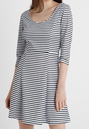 Femme portant une robe blanche à rayures horizontales noires, manches trois-quarts et encolure dégagée, debout devant un fond uni.