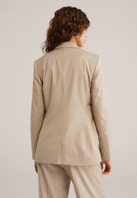 Smėlio spalvos švarkas su siaurais dryžiais, pasiūtas pagal individualų modelį, turintis struktūruotus pečius ir lygią tekstūrą, su vienu skeltuku gale.