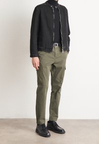 Černá džínová bunda, olivové cargo kalhoty a černé boty. Bunda má cvočky a zapínání na zip; kalhoty mají boční kapsy.