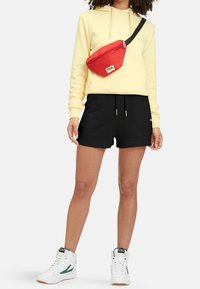Sweat à capuche jaune, short noir et baskets blanches avec des accents verts. Sac bandoulière rouge avec logo. Tenue décontractée sportive.