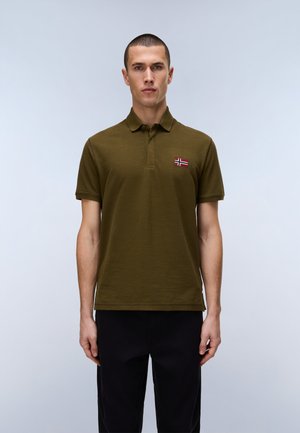 Uomo che indossa una polo a maniche corte verde oliva con una toppa della bandiera norvegese, in piedi davanti a uno sfondo grigio chiaro uniforme.