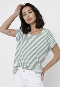 T-shirt à manches courtes vert et blanc rayé avec un col rond et une coupe décontractée, assorti à un jean blanc et des accents dorés minimalistes.