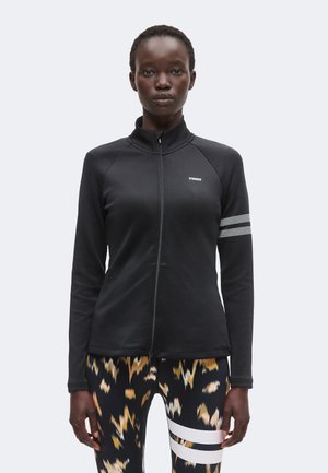 Kvinde iført en sort lynlåstrøje med hvide striber på det ene ærme og mønstrede leggings med hvide striber på det ene lår.