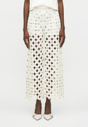 Weiße, hoch taillierte, weit geschnittene Häkelhose mit Blumenmustern und kleinen metallischen Akzenten, getragen zu weißen spitz zulaufenden Buckle-Pumps.