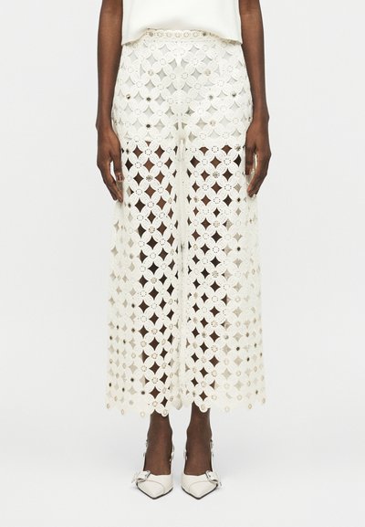 Pantalon blanc à taille haute et jambes larges en crochet avec des motifs floraux et de petits accents métalliques, porté avec des talons blancs pointus à boucle.