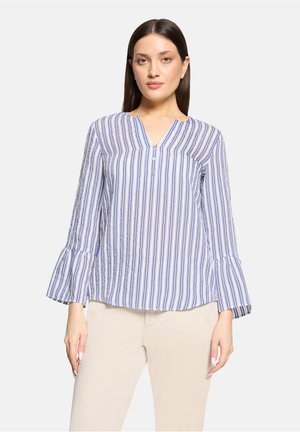 Femme portant un chemisier à rayures verticales bleu et blanc avec des manches évasées et un pantalon beige, debout devant un fond clair uni.