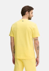 T-shirt di cotone gialla con maniche corte e collo rotondo. Presenta un piccolo logo verde sulla parte posteriore. Tessuto morbido, vestibilità rilassata.