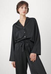 Svart satin pyjamas-set bestående av en knäppbar topp med krage och långa ärmar, tillsammans med byxor med snörning och vida ben. Slät struktur.