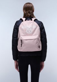 Zaino di tessuto rosa con un logo centrale, spallacci regolabili, una tasca anteriore con cerniere e una texture liscia, indossato su un giubbotto imbottito nero.