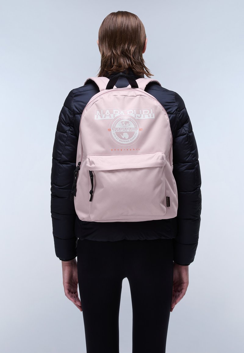 Zaino di tessuto rosa con un logo centrale, spallacci regolabili, una tasca anteriore con cerniere e una texture liscia, indossato su un giubbotto imbottito nero.