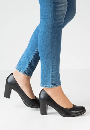 Sorte højhælede pumps med en glat læderlignende overflade, rund tå-design og blokhæle. Båret med tætsiddende blå jeans.