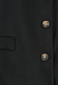 Tumma kangas, jossa on kaksi pyöreää, marmoroitua ruskeaa nappia ja vaakasuora kenkätasku kangaslappu vasemmalla puolella.