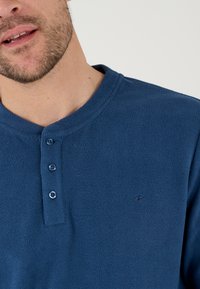 Blauwe fleece henley-shirt met een ronde kraag, drie knoopsluitingen en een subtiel geborduurd logo aan de linkerkant. Zachte textuur.