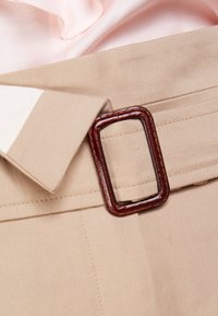 Un tissu beige avec une texture lisse présente une boucle de ceinture rectangulaire marron. Un tissu satin rose clair est partiellement visible à l'arrière-plan.