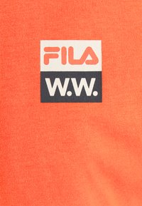 Korálové tričko Fila s texturovanou tkaninou. Na přední straně se nachází tkaná nášivka v bílé a námořnické barvě s nápisem "FILA" nad nápisem "W.W." vyvedeným tučným písmem.