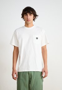 White cotton t-shirt with short sleeves, featuring a black heart-shaped logo on the left chest pocket. Paired with green pants.