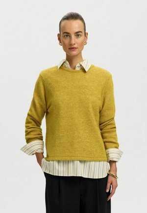 Woman with hair tied back wearing gold hoop earrings, mustard sweater over striped shirt, black pants, and gold bracelet against plain background.