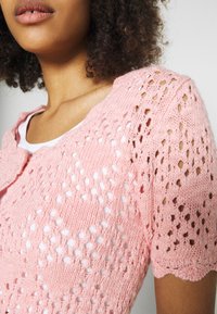 Rosa gestrickte Strickjacke mit kurzen Ärmeln, versehen mit einem Durchbruchsdesign und gewellten Kanten, übereinander getragen mit einem weißen Oberteil.