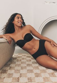 Czarny biustonosz bez ramiączek i dopasowane, wysokie majtki bikini. Gładki materiał, prosty wzór, bez nadruków. Modelka leży na beżowej sofie.