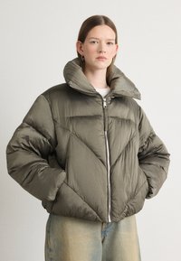 Pufferjakke i olivengrønn med en glanset finish, med høy krage, glidelås og skrå quiltet mønster. Brukt sammen med lys denim.