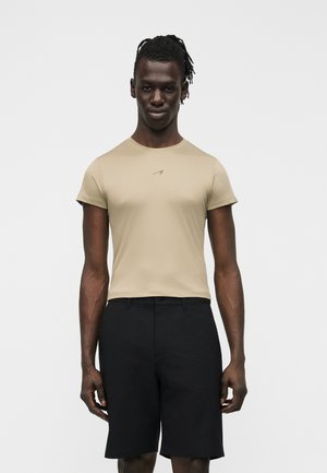 Moški s temno poltjo in dredži, oblečen v bež oprijeto majico s kratkimi rokavi in črne kratke hlače do kolen, stoji pred enakomerno belo podlago.